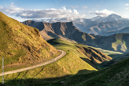 Fotografi Mountain valley shined with the sunset sun with the road on an abrupt slope