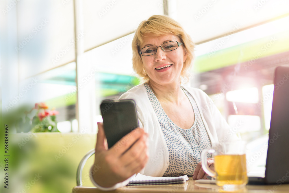 A middle-aged woman with a laptop works in a cafe in the office, she is a freelancer. A woman with glasses sits at a table with a Cup of herbal tea, looks on camera and smiles. A middle-aged woman with a laptop works in a cafe in the office, she is a freelancer. A woman with glasses sits at a table with a Cup of herbal tea, looks on camera and smiles.