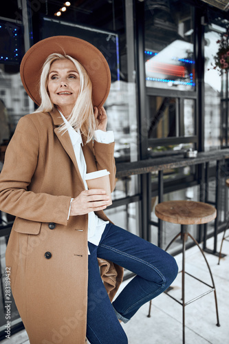 Gladsome adult woman in coat and hat smiling at the cafe terrace