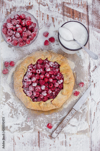 Homemade fruit pie (galette) made with fresh raspberries with powdered sugar on wooden table. Open pie, raspberry tart. Summer berry dessert. Flat lay.