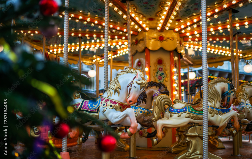 Сarousel horses to traditional Christmas market. Outdoor fair on winter day. View through the frost pine branch. Happy new year. Festive street decor in winter holidays.