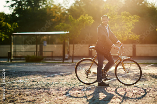 Fototapeta Naklejka Na Ścianę i Meble -  Bearded Businessman in business suit riding on retro bicycle to work on urban street in the morning on sunset