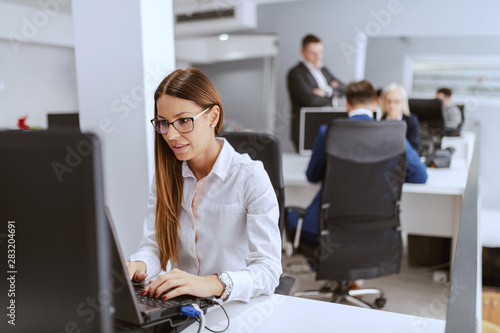 Dedicated focused highly motivated businesswoman sitting at workplace and using PC. hands on keyboard. In background her colleagues working.