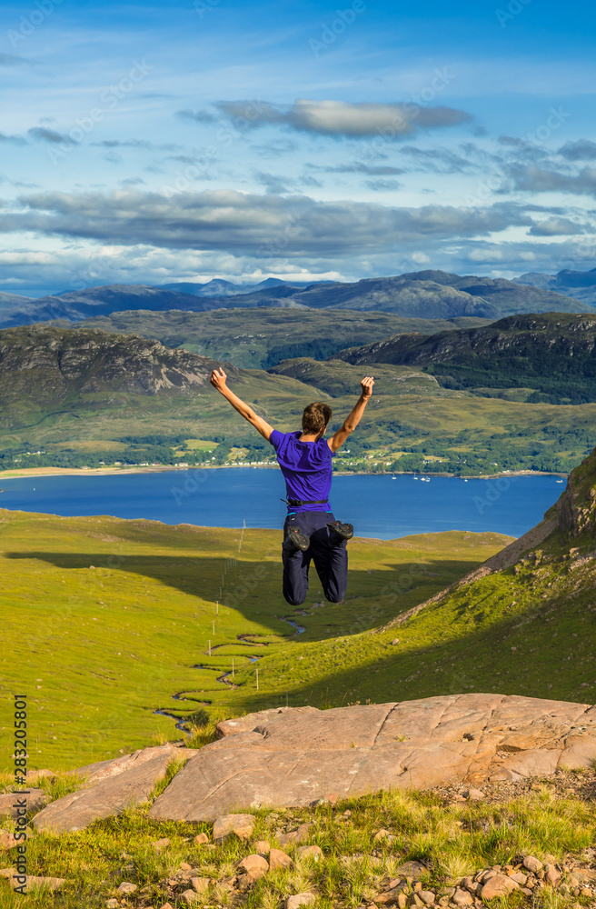 Young Boy Jumps Happily On Rock In Front Of Spectacular Landscape Of Applecross Pass In Scotland