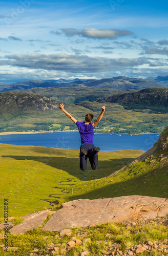 Young Boy Jumps Happily On Rock In Front Of Spectacular Landscape Of Applecross Pass In Scotland