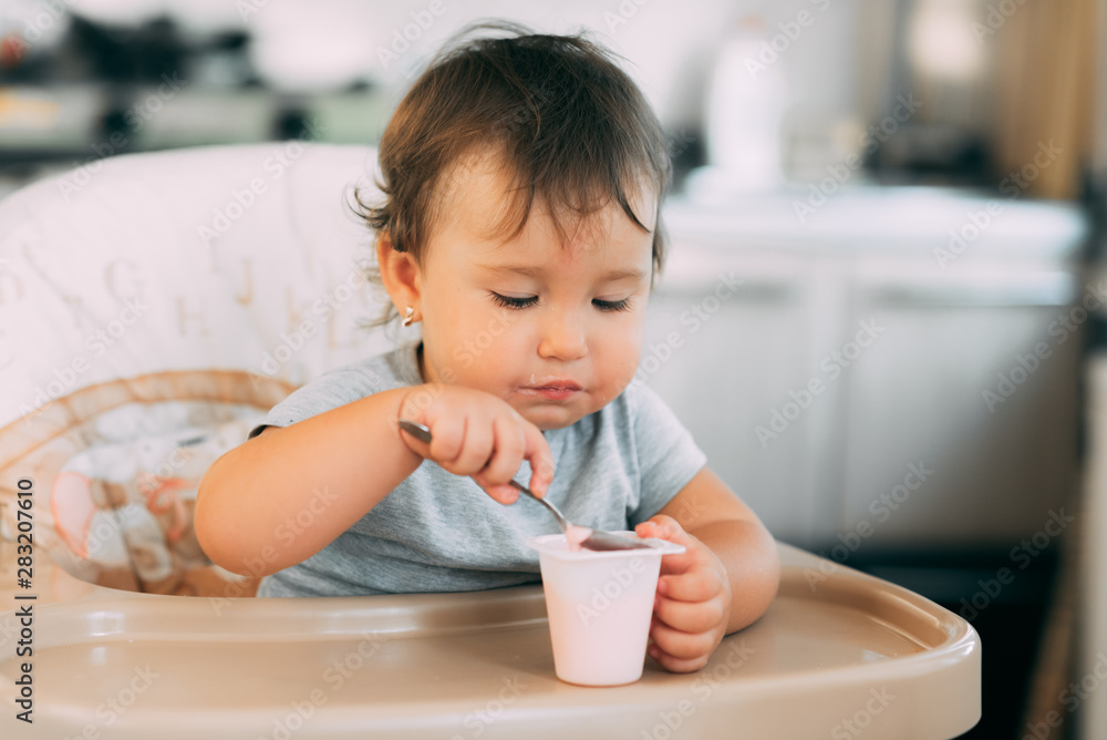 Cute, little girl eating yogurt in the kitchen during the day in a high chair Cute, little girl eating yogurt in the kitchen during the day in a high chair