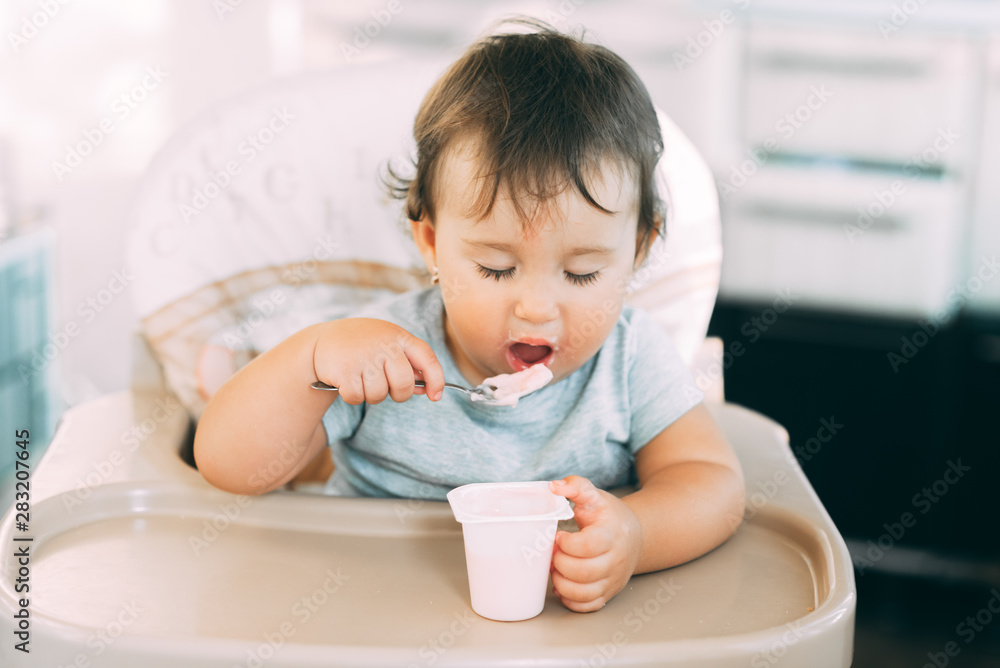 Cute, little girl eating yogurt in the kitchen during the day in a high chair Cute, little girl eating yogurt in the kitchen during the day in a high chair