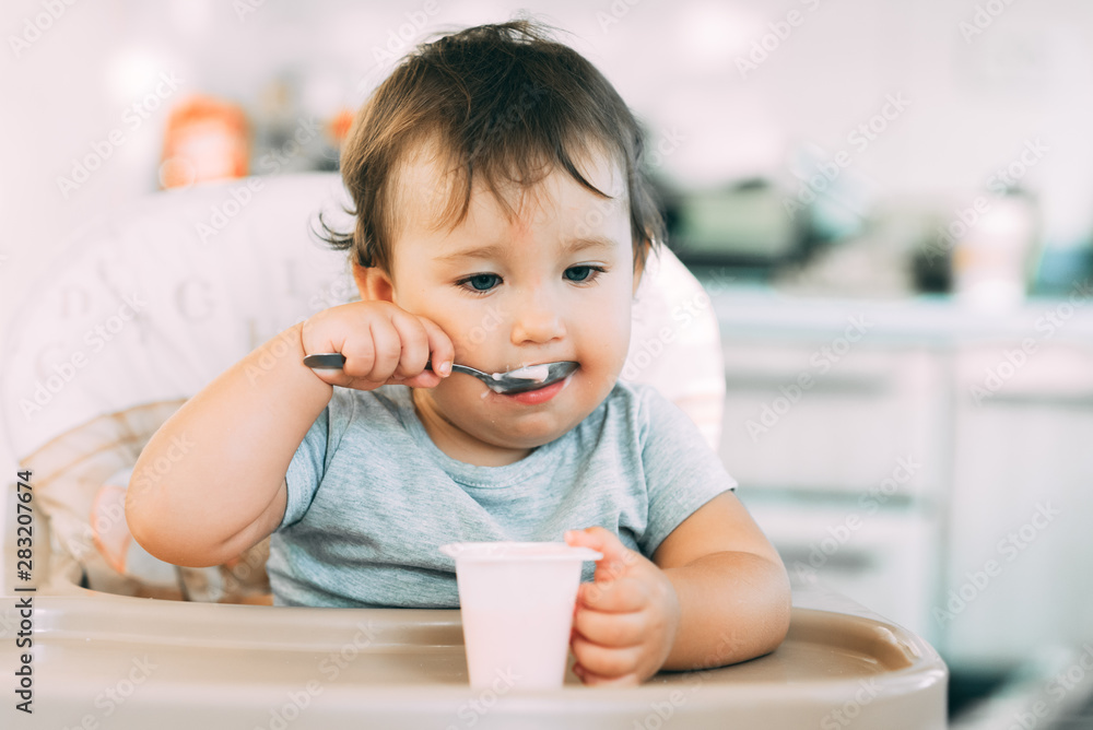 Cute, little girl eating yogurt in the kitchen during the day in a high chair Cute, little girl eating yogurt in the kitchen during the day in a high chair