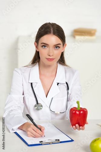 front view of dietitian holding bell pepper and writing in clipboard at workplace