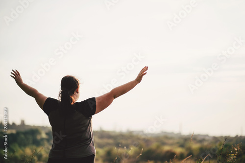 Body positive, freedom, high self esteem, confidence, happiness, inspiration, success, positive affirmation. Overweight woman celebrating rising hands to the sky on summer meadow.