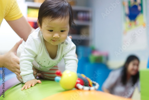 Young little Asian cute baby crawling on circular cushion practicing balance the body teaching by her preschool teacher in gym. The kid looking at toy and trying to catch it. Play and learn concept.