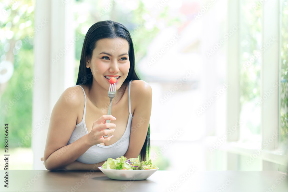 Young beautiful Asian girl eating green salad sitting on the wooden table with smiley face and feeling happy. She holding fork with tomato looking out the window. Diets and healthy food concept, Young beautiful Asian girl eating green salad sitting on the wooden table with smiley face and feeling happy. She holding fork with tomato looking out the window. Diets and healthy food concept,