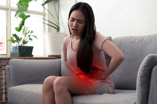 Young cute Asian girl having stomachache sickness problem sitting on sofa at home with painful feeling. She holding her belly and twisting the body with both hands. Red mark shows area of the pain.