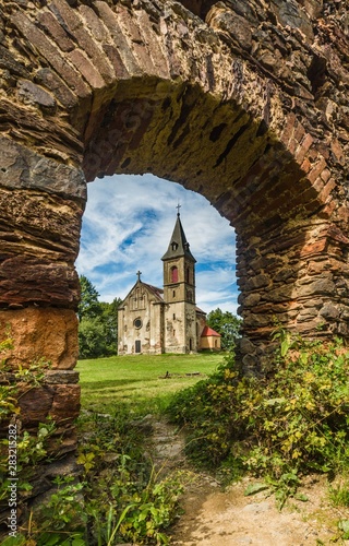 Krasikov, Kokasice / Czech Republic - August 9 2019: View of the church of Mary Magdalena through a door in a stone wall. Sunny day. Green grass and blue sky. Vertical image.