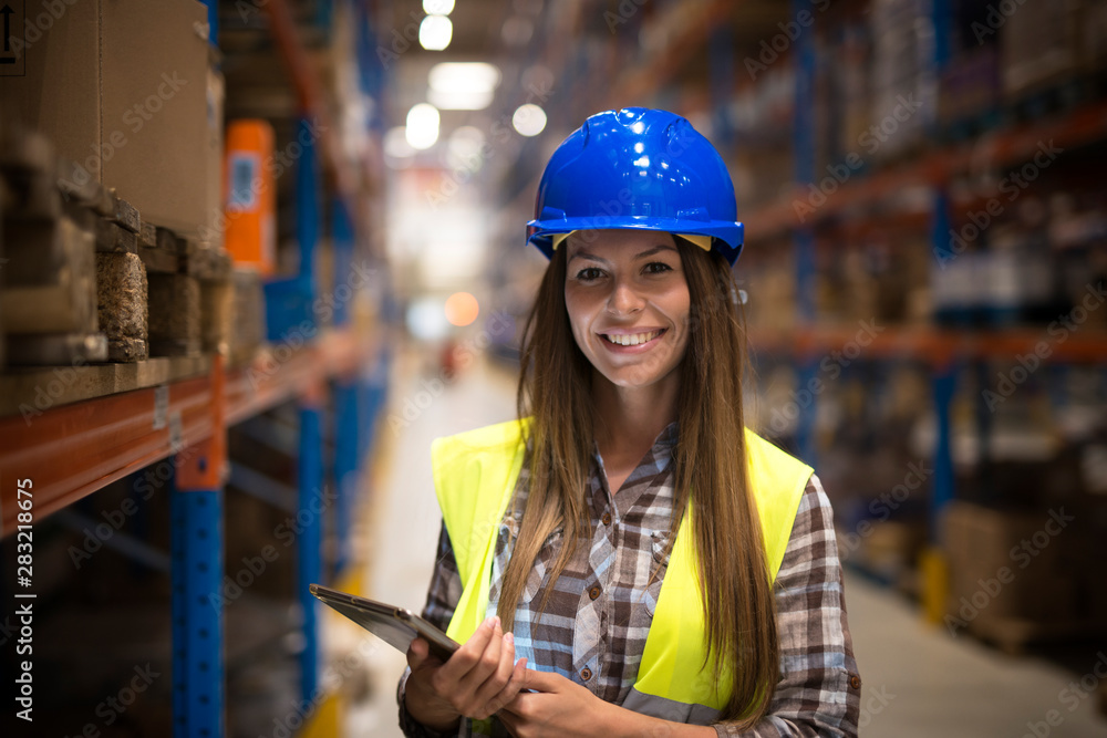 Portrait of smiling woman in protective uniform with hardhat holding tablet in warehouse center. - obrazy, fototapety, plakaty 