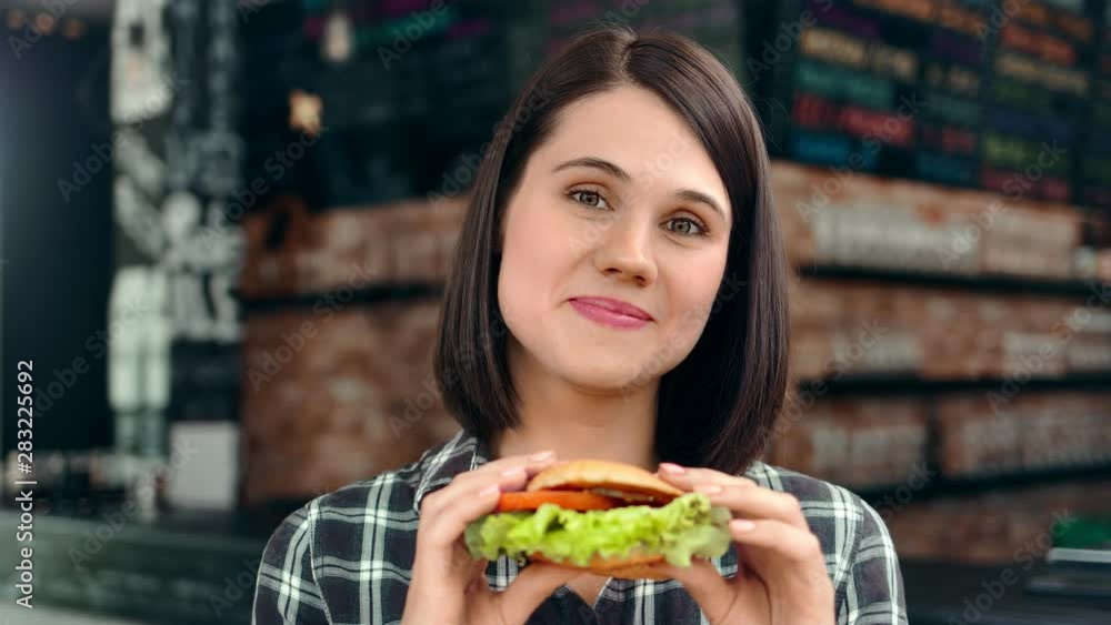 Beautiful face of friendly young girl posing holding fresh juicy burger looking at camera Beautiful face of friendly young girl posing holding fresh juicy burger looking at camera