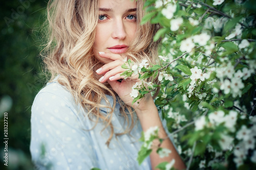 Beautiful happy young woman enjoying smell in a flowering spring garden. Blonde with blue eyes and blue dress.