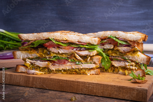 Delicious sandwich cut into triangles - toasted bread with chicken breast, fried bacon, vegetables, pickles with mustard and arugula closeup on a dark background, copy space
