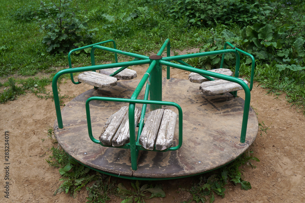 Small children's old carousel in a courtyard. Green knobs, shabby wooden seats, floor with peeling paint. Ground around, green grass in the background Small children's old carousel in a courtyard. Green knobs, shabby wooden seats, floor with peeling paint. Ground around, green grass in the background