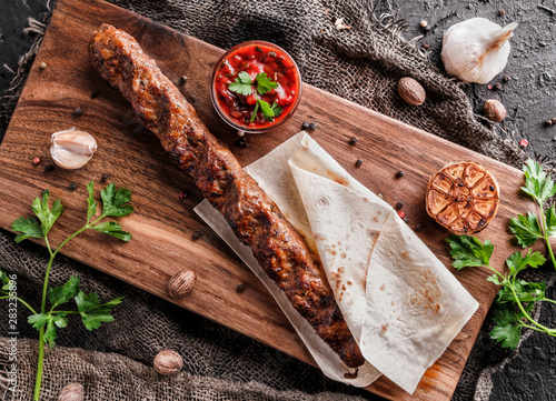 Grilled meat kebab in pita bread with tomato sauce and herbs on wooden board over dark background. Top view