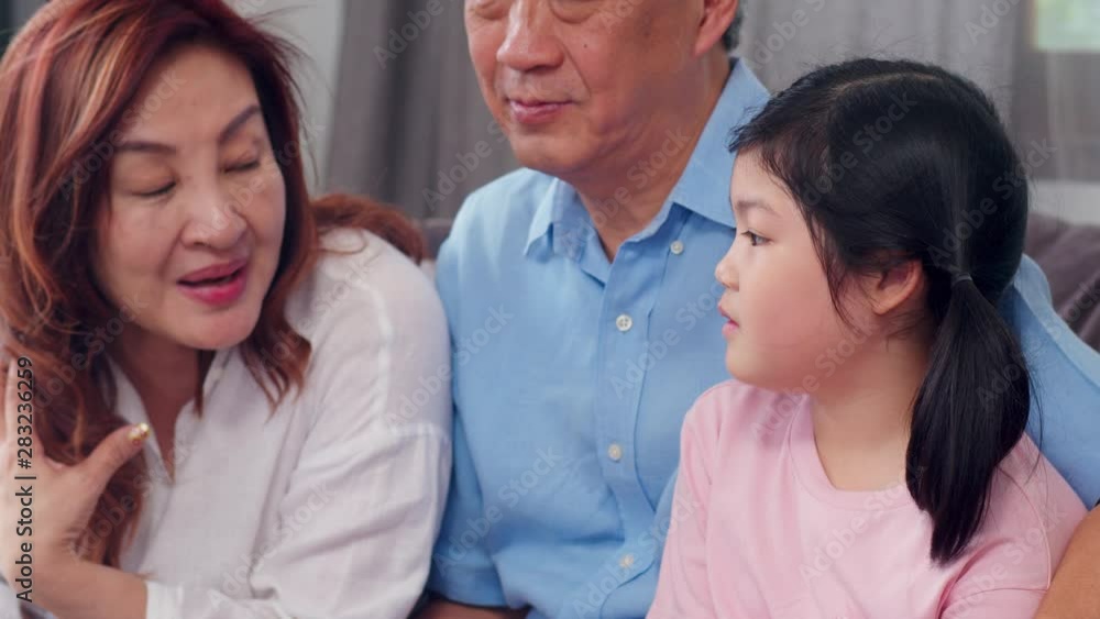 Asian grandparents talking with granddaughter at home. Senior Chinese, old generation, grandfather and grandmother family time relax with young girl kid on sofa in living room concept. Slow motion.