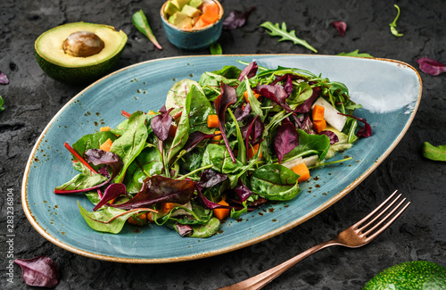 Fresh green salad with mixed greens, spinach arugula, pumpkin, avocado in plate over dark background. Healthy food, clean eating, vegan dieting, top view