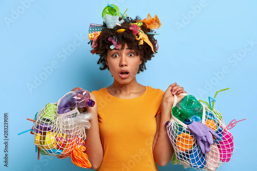 Ecology and environment concept. Emotional dark skinned woman supports litter reduction  collects garbage in net bags  puzzled with plastic pollution  being ecologically friendly  stands indoor