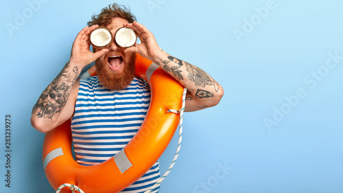 Funny redhead holiday maker wears sailor vest, poses with inflated lifebuoy, ready to give help for people at sea, holds coconuts on eyes, pretends looking in binocular notices something into distance