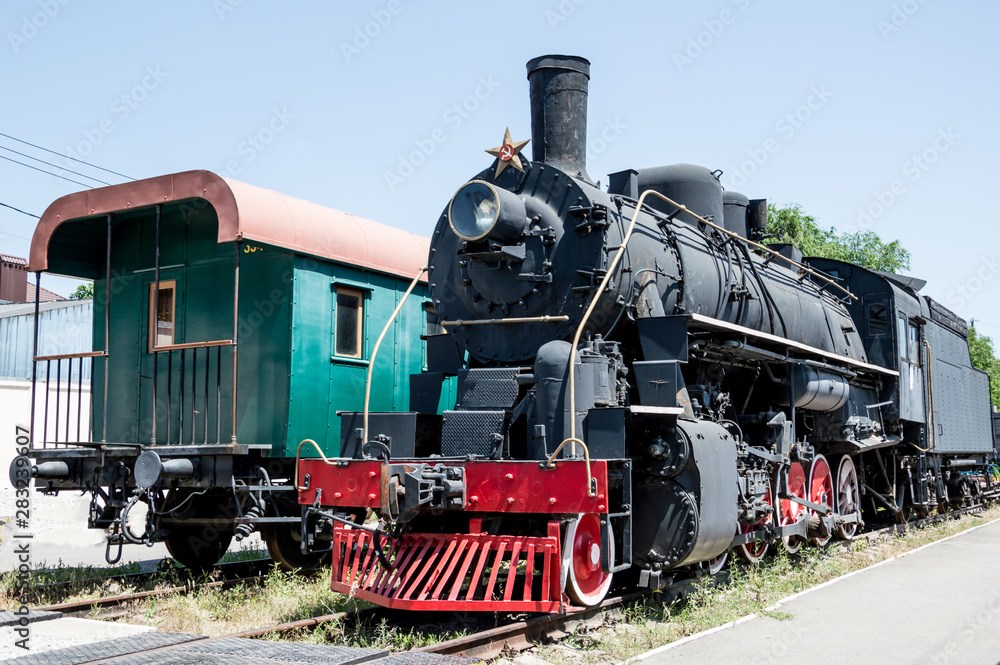 Fototapeta premium A black Soviet steam locomotive with a star and an old passenger green wooden carriage stand nearby on rails.