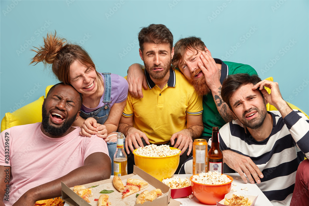 Five fellows laugh loudly as watch funny comedy movie or comic show, enjoy hilarious scene, eat delicious snack, pose at sofa near small table with fast food, spend weekend or free time together.