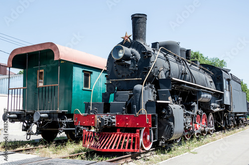 A black Soviet steam locomotive with a star and an old passenger green wooden carriage stand nearby on rails.