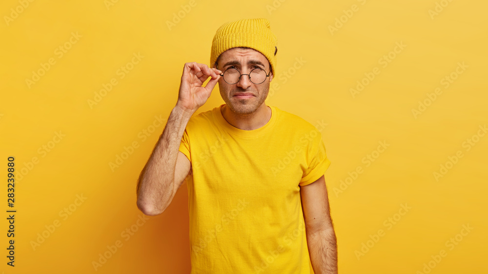 Discontent male model looks thoroughly through round glasses, has bad eyesight, tries to notice something little, touches frame of spectacles, has attentive look, dressed in casual yellow outfit