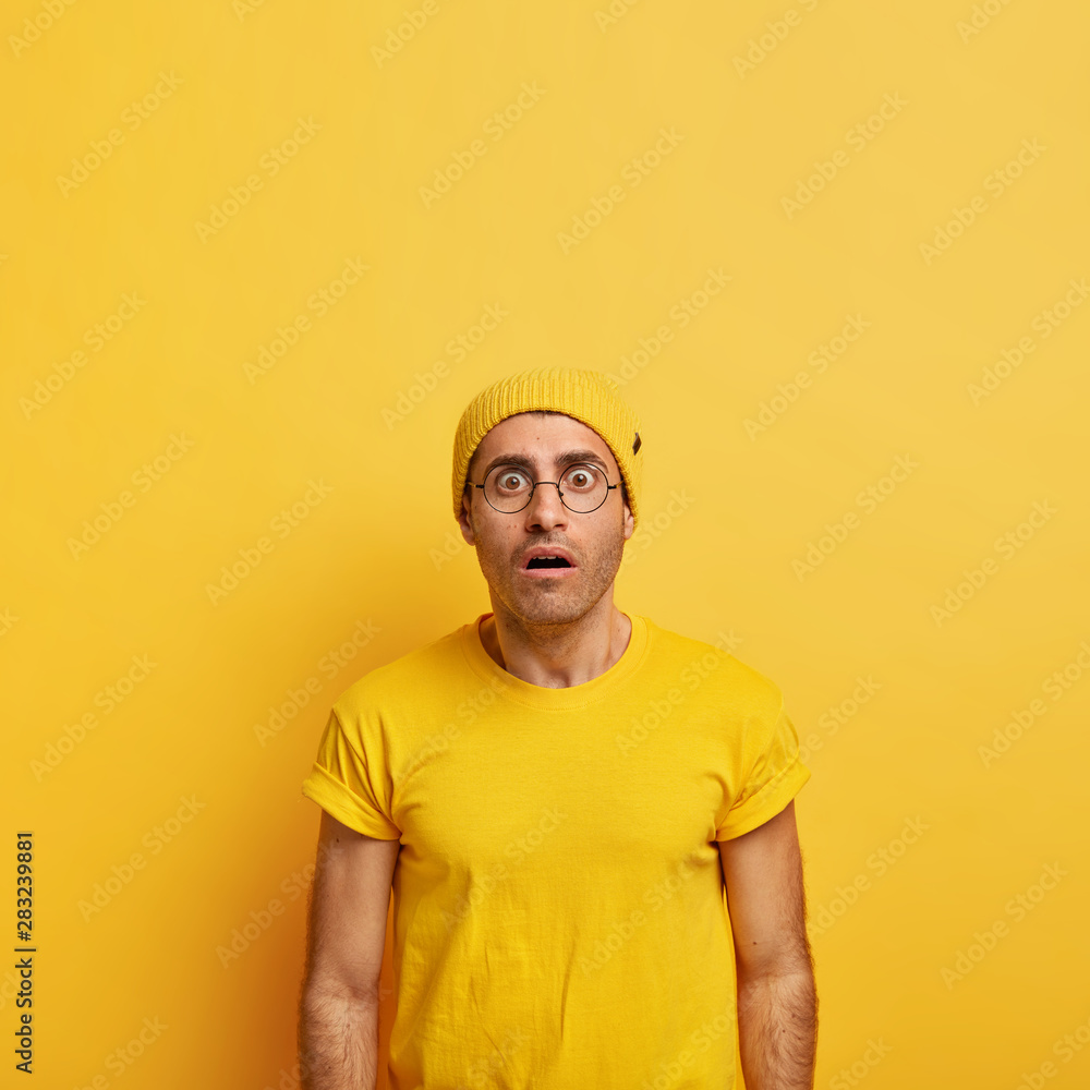 Intense stupefied man stares at camera with bugged eyes, cannot believe in bad news, realizes he made huge mistake, stunned by something, wears bright yellow hat and casual t shirt. Omg concept