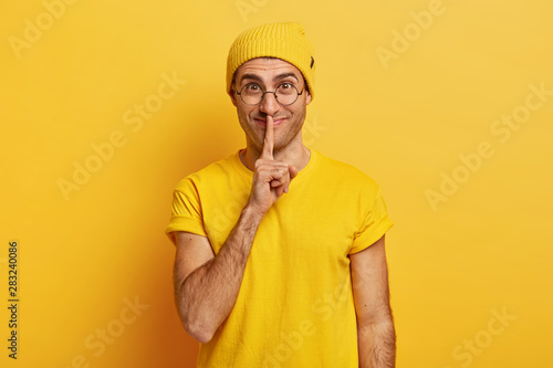 Mysterious satisfied man asks stay silent, shows shush gesture, keeps index finger over lips, has delighted face expression, shares secret with girlfriend, wears yellow t shirt, stylish hat.