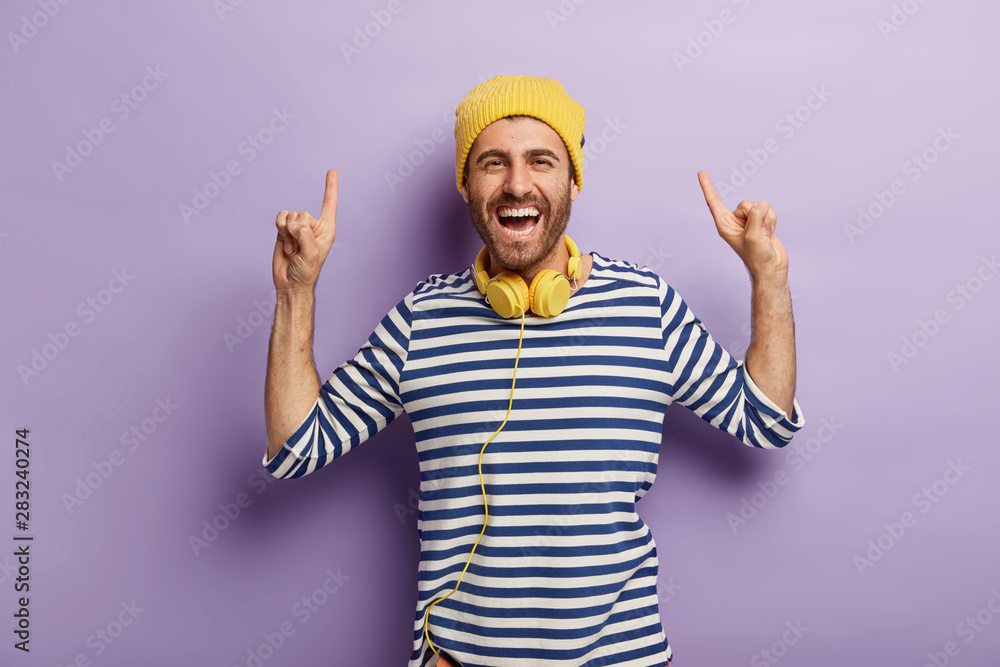 Cheerful unshaven man prefers going upwards, raises index fingers above, wears yellow hat and striped jumper, listen music with headphones during spare time, stands against purple background