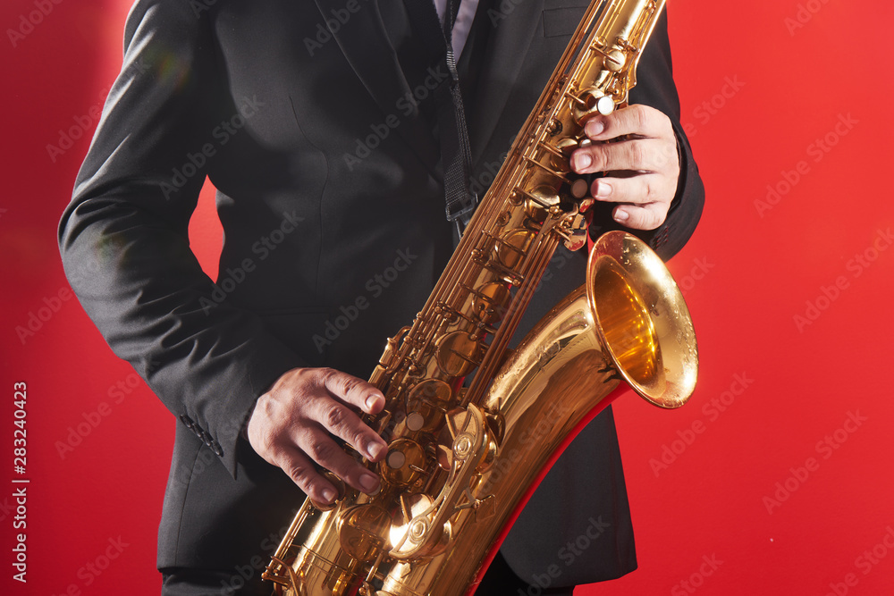 Obraz premium Portrait of professional musician saxophonist man in suit plays jazz music on saxophone, red background in a photo studio