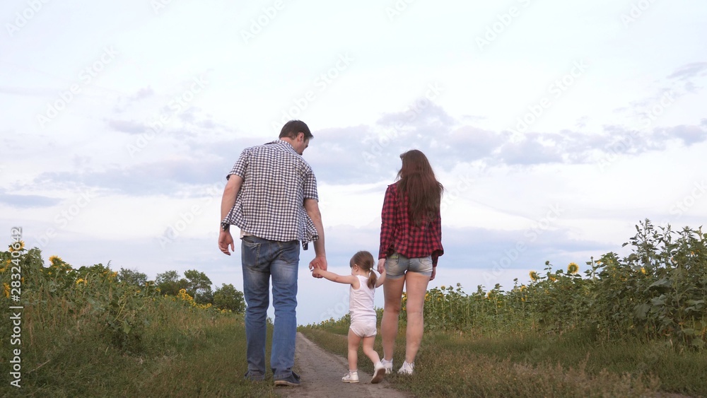 Family with small child walks along road and laughs next to field of sunflowers. Child is riding in arms of his father and mother. Mom, dad and daughter are resting together outside city in nature