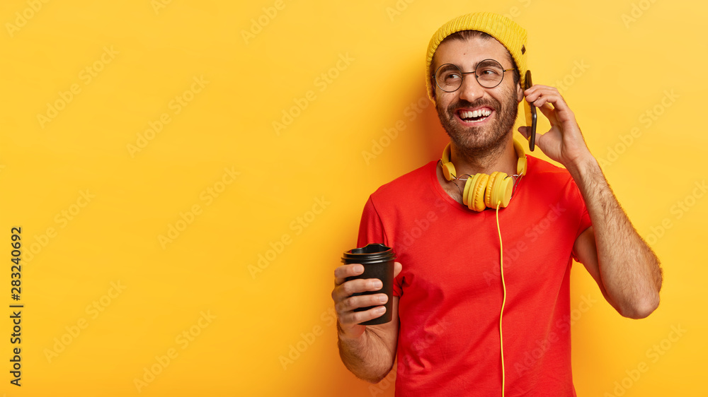 Indoor shot of cheerful millennial man speaks on mobile phone, looks away, holds takeout coffee makes call to friend dressed in stylish yellow hat red t shirt, enjoys spare time. People, communication Indoor shot of cheerful millennial man speaks on mobile phone, looks away, holds takeout coffee makes call to friend dressed in stylish yellow hat red t shirt, enjoys spare time. People, communication