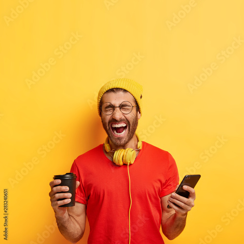 Overemotive positive man laughs from funny content publication, poses indoors with smartphone and disposable cup of coffee, keeps mouth wide opened, wears bright vivid clothes, isolated on yellow wall © Wayhome Studio