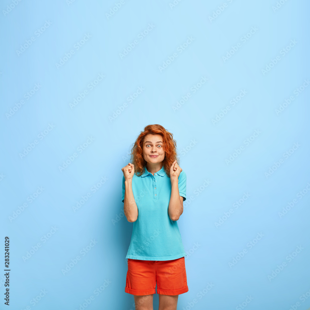 Fototapeta premium Hopeful ginger female model clenches fists with big desire to achieve goal, looks with intriguing expression, wears casual blue t shirt, red shorts, shows victory gesture, cannot wait for something