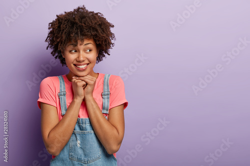 Curly haired good looking girl keeps hands together, looks away with gentle smile, feels touched to receive lovely present, poses indoor against purple background, free space for your promotion