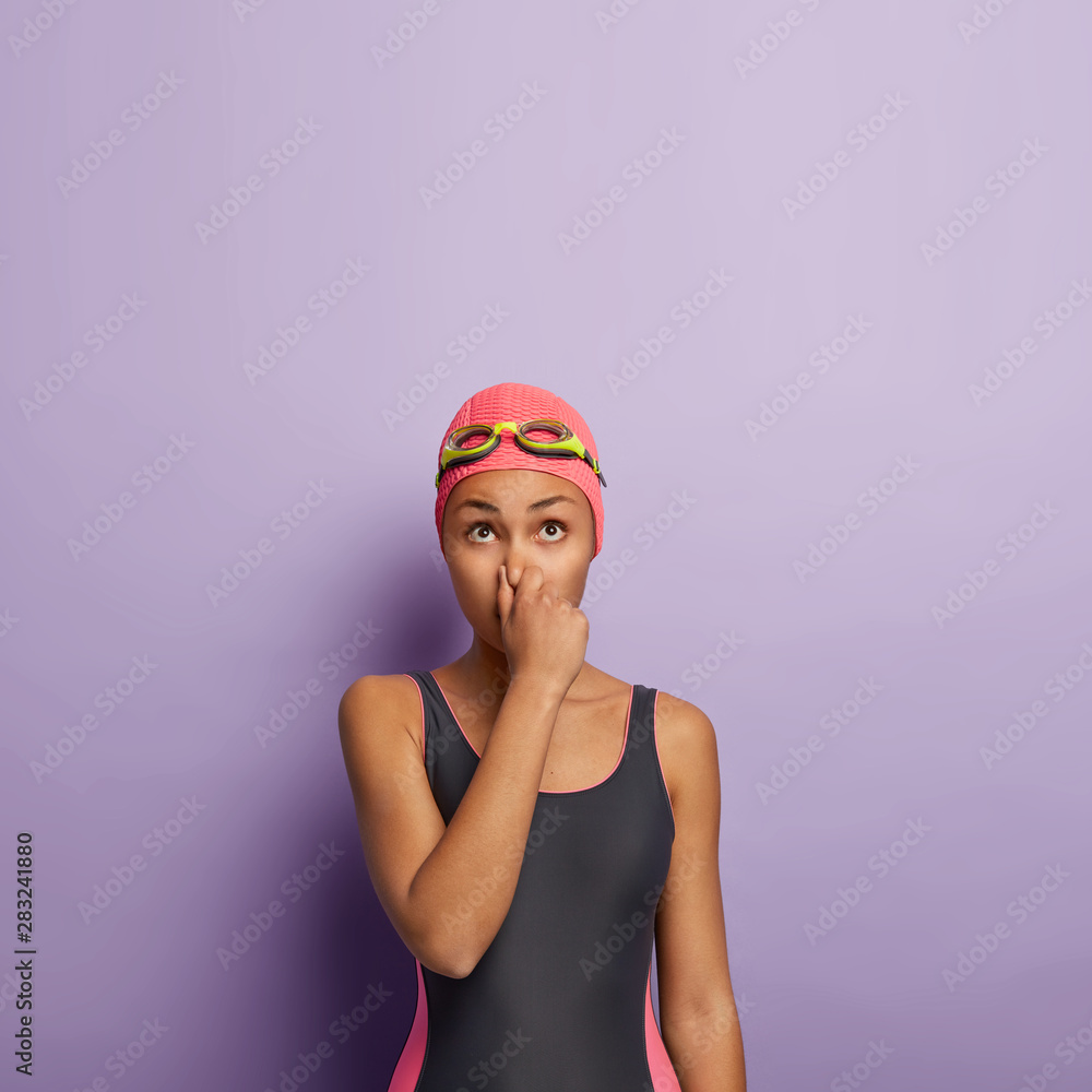 Vertical shot of slim woman holds breath, covers nose, preapres for diving under water, looks above, wears pink swimcap, black swimsuit, goggles, spends free time in swimming pool of fitness center Vertical shot of slim woman holds breath, covers nose, preapres for diving under water, looks above, wears pink swimcap, black swimsuit, goggles, spends free time in swimming pool of fitness center