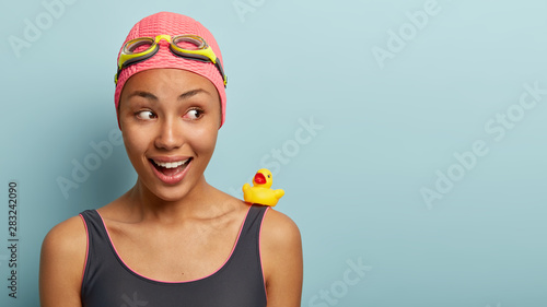 Headshot of charming dark skinned female swimmer wears pink bathing cap, swimming glasses, swimsuit, focused aside, swims with rubber duck, rejoices good vacation. People and lifestyle concept