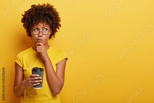 Pensive displeased woman with dark skin, holds chin and looks away, contemplates about something, wears yellow t shirt, holds disposable cup of coffee, smirks face and being upset by bad thoughts