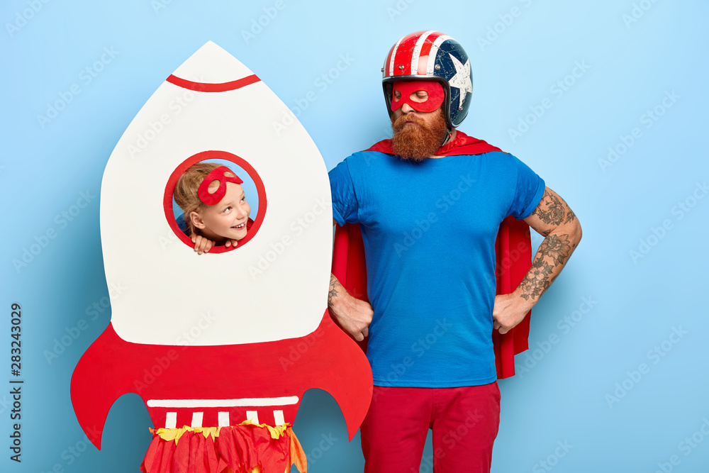 Obraz premium Serious father keeps hands on waist, wears headgear, blue shirt, red cloak looks at daughter play game together have fun at costume party with decoration. Lovely kid looks through rocket window at dad