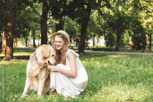 beautiful girl in white dress and straw hat petting golden retriever while sitting on meadow