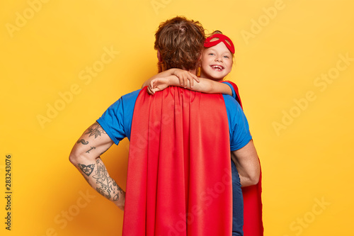 Affectionate daddy wears red cape, stands back to camera, carries small child who embraces him and smiles happily, dressed in superhero costumes, isolated on yellow background, have fun together