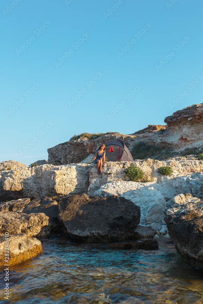 Girl in a dress with a tent on the sea stone shore. View from the water