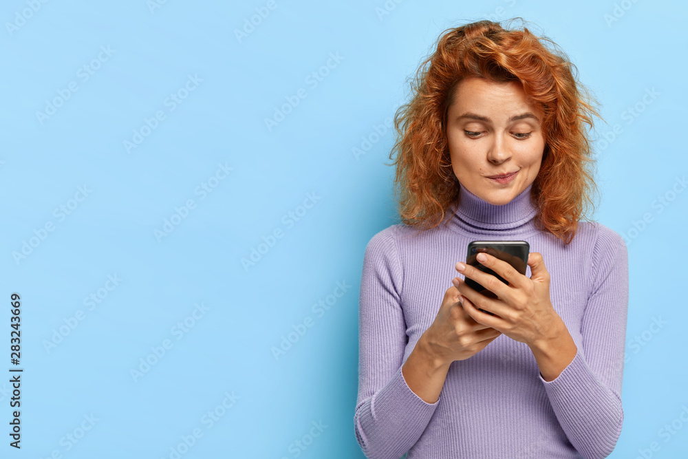 Portrait of pleasant looking redhead lady focused in smartphone device, checks email box, chats online, gets notification, connected to wireless internet, wears casual sweater, models indoor Portrait of pleasant looking redhead lady focused in smartphone device, checks email box, chats online, gets notification, connected to wireless internet, wears casual sweater, models indoor