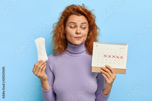 Photo of young woman looks at period calendar, checks menstruation days, holds soft sanitary pad, puzzled with delay of periods, has premesntrual syndrom as intense mood swings, prepares for PMS